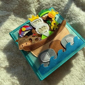 May include: A felt book with a teal cover featuring a brown ark filled with felt animals and people. The animals include a giraffe, elephant, and cow. The people are wearing crowns and hats. Blue felt waves are at the bottom.