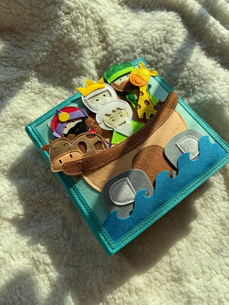 May include: A felt book with a teal cover featuring a brown ark filled with felt animals and people. The animals include a giraffe, elephant, and cow. The people are wearing crowns and hats. Blue felt waves are at the bottom.