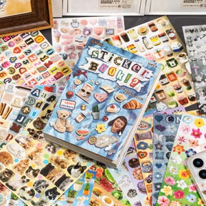 Puede incluir: Un libro de pegatinas con una cubierta de cuadr&iacute;cula azul que presenta el texto Sticker Book y varias ilustraciones peque&ntilde;as que incluyen un oso de peluche, un cruas&aacute;n y un gato. El libro est&aacute; rodeado por varias hojas de pegatinas coloridas.