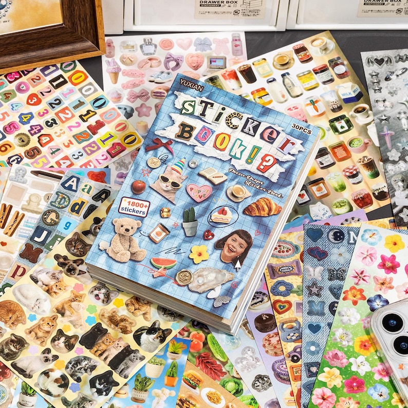 Puede incluir: Un libro de pegatinas con una cubierta de cuadr&iacute;cula azul que presenta el texto Sticker Book y varias ilustraciones peque&ntilde;as que incluyen un oso de peluche, un cruas&aacute;n y un gato. El libro est&aacute; rodeado por varias hojas de pegatinas coloridas.