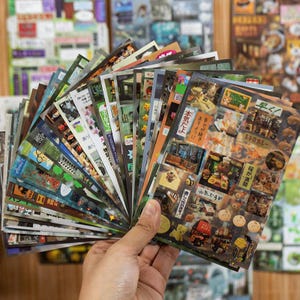 May include: A hand holds a fan of colorful stickers with various designs, including Japanese text, illustrations of food, and other decorative elements. The stickers are in various shapes and sizes, with a mix of colors.