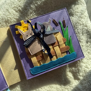 May include: A handmade felt quiet book page featuring a scene with two animal figures in a woven basket on a purple background. The scene includes a yellow sun, blue water, and green grass. A navy blue ribbon with white polka dots is tied around the basket.