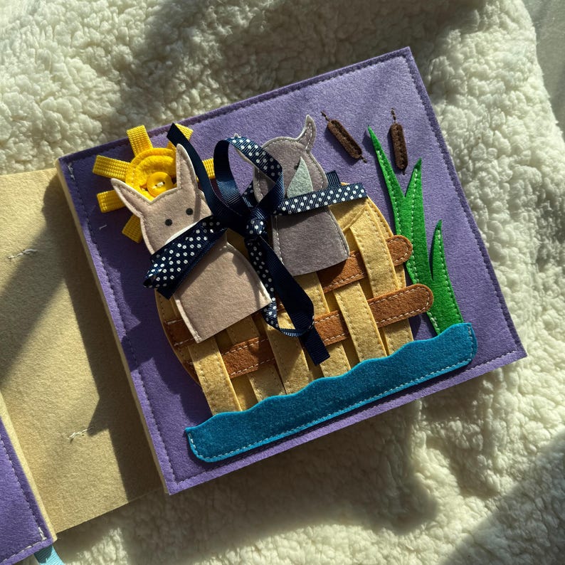 May include: A handmade felt quiet book page featuring a scene with two animal figures in a woven basket on a purple background. The scene includes a yellow sun, blue water, and green grass. A navy blue ribbon with white polka dots is tied around the basket.