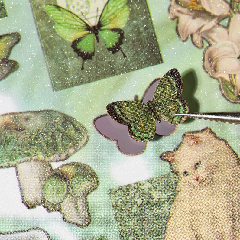May include: Close-up of various stickers with a whimsical theme. The stickers include green butterflies, mushrooms, and a white cat. The stickers have a glittery finish and are set against a light green background.