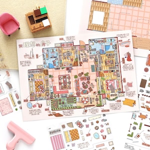 May include: A printed paper sheet featuring a detailed top-down floor plan illustration titled in Chinese characters, surrounded by smaller cut-out furniture icons, a pink miniature chair, and a wooden cabinet model on a light surface.