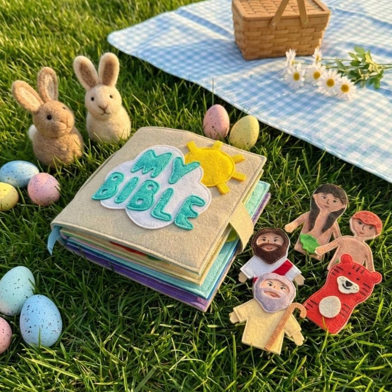 May include: A felt book titled "MY BIBLE" with a cloud and sun design, surrounded by felt figures and Easter eggs. Two bunny figures and a picnic basket are also present, all set on a grassy surface.