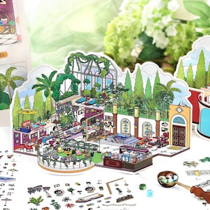 Op de afbeelding: Een kleurrijk, gedetailleerd papieren diorama van een caféscène, met een kas, zitplaatsen buiten en een bar. De illustratie bevat verschillende planten, meubels en decoratieve elementen. Kleine accessoire stickers liggen verspreid.