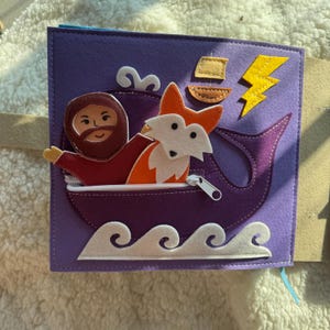 May include: A felt quiet book page featuring a purple boat with a zipper, holding a cartoon character and a fox. The scene includes white waves, a yellow lightning bolt, and a small boat. The background is a soft, off-white fabric.