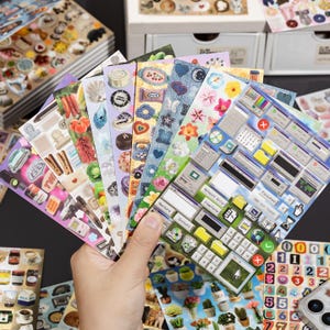 Puede incluir: Un abanico de varias hojas de pegatinas con dise&ntilde;os diversos que incluyen ventanas de interfaz de computadora retro, patrones florales, texturas de mezclilla, alimentos y varios objetos peque&ntilde;os, dispuestos en capas.