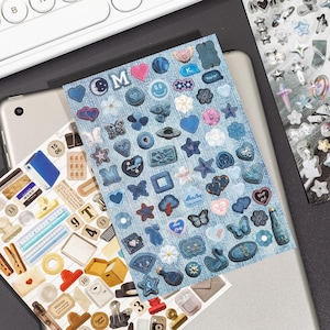 Puede incluir: Una hoja de pegatinas rectangular con un fondo de textura vaquera, cubierta con varios iconos adhesivos peque&ntilde;os en tonos azules, incluyendo corazones, estrellas, mariposas, flores, caras sonrientes y burbujas de texto con las letras M y K.