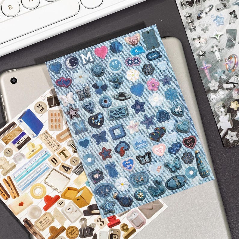 Puede incluir: Una hoja de pegatinas rectangular con un fondo de textura vaquera, cubierta con varios iconos adhesivos peque&ntilde;os en tonos azules, incluyendo corazones, estrellas, mariposas, flores, caras sonrientes y burbujas de texto con las letras M y K.