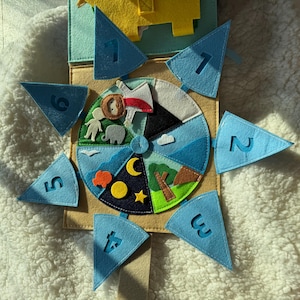 May include: A felt educational toy featuring a circular design with various scenes and blue triangular pieces numbered 1-6. The center depicts day and night scenes with animals. The toy is made of various colors of felt.