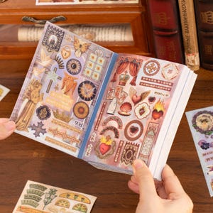 May include: A sticker book with various vintage-style designs, including crowns, hearts, and celestial elements. The stickers feature gold, red, and blue accents, with some text in a foreign language. The book is open, revealing multiple pages of stickers.