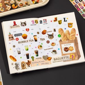 Puede incluir: Una p&aacute;gina de agenda abierta para julio de 2026 con un calendario de cuadr&iacute;cula decorado con pegatinas de gatos, perros, t&eacute; de burbujas, caf&eacute; y pan. La p&aacute;gina incluye textos como 2026, JUL, BUBBLE TEA, TEA, BAGUETTE y una To-Do List.