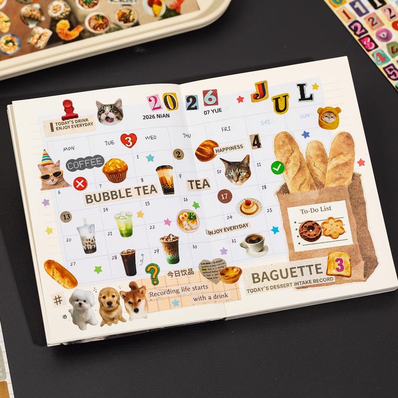 Puede incluir: Una p&aacute;gina de agenda abierta para julio de 2026 con un calendario de cuadr&iacute;cula decorado con pegatinas de gatos, perros, t&eacute; de burbujas, caf&eacute; y pan. La p&aacute;gina incluye textos como 2026, JUL, BUBBLE TEA, TEA, BAGUETTE y una To-Do List.