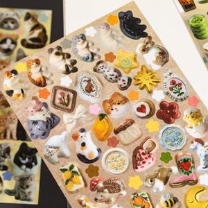 Puede incluir: Una hoja de pegatinas con una variedad de adhesivos tridimensionales que incluyen gatos, conejos, flores, frutas, estrellas, corazones y objetos peque&ntilde;os como una bota y un hongo, dispuestos sobre una hoja de soporte rectangular de color marr&oacute;n claro.