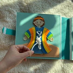 May include: A felt activity book with a blue cover and a character wearing a colorful robe. The character has a lace-up design on the front. The book is open, and a hand is shown holding the white lace.