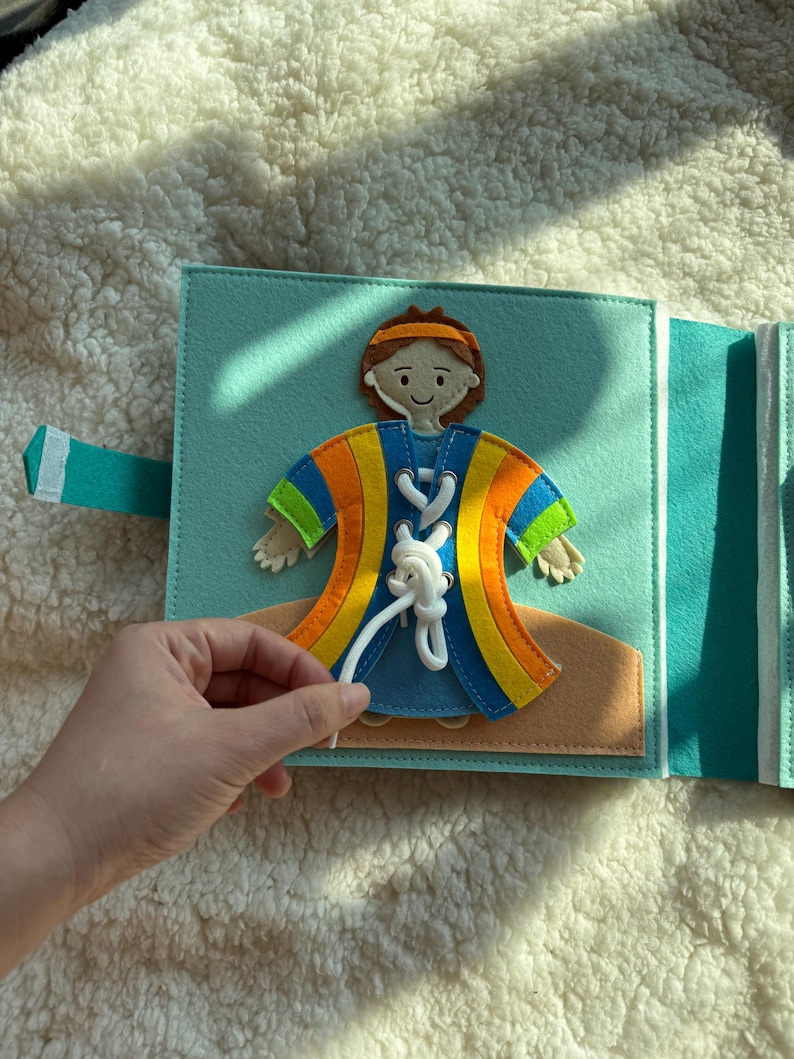 May include: A felt activity book with a blue cover and a character wearing a colorful robe. The character has a lace-up design on the front. The book is open, and a hand is shown holding the white lace.