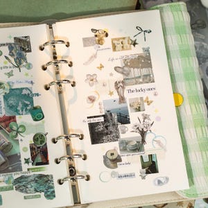 May include: Open planner with decorative stickers, including floral, angel, and butterfly designs. The text "The lucky ones" and "Life is not all roses" are visible. The planner has silver rings and a green and white checkered cover.
