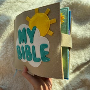 May include: A tan felt book with the words "MY BIBLE" in teal letters on a white cloud. A yellow sun with yellow rays is stitched on the cover. The book is secured with a tan strap.