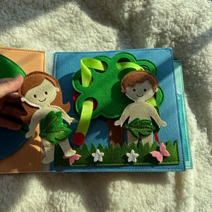 May include: A felt quiet book page featuring a tree with red apples, two figures, and a garden scene. The figures have brown hair and are adorned with leaf coverings. The scene includes green grass, white flowers, and pink butterflies.