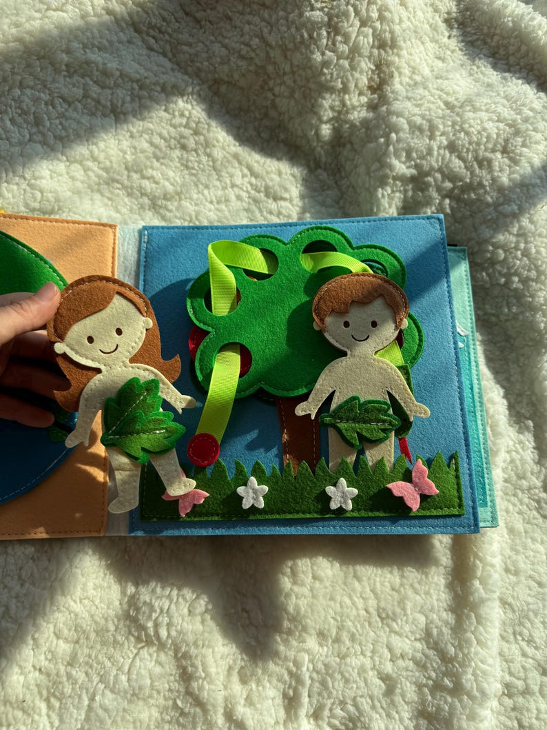 May include: A felt quiet book page featuring a tree with red apples, two figures, and a garden scene. The figures have brown hair and are adorned with leaf coverings. The scene includes green grass, white flowers, and pink butterflies.