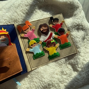 May include: A felt book with colorful figures of Jesus and children. The figures are on a beige background, with a blue book cover on the left. The figures have various skin tones and hair colors.