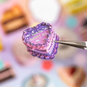 Puede incluir: Un pastel en miniatura con forma de coraz&oacute;n con glaseado morado y rosa, que presenta el texto "Not me turning 24" en letras blancas. El pastel est&aacute; sujeto por unas pinzas de metal plateado y decorado con peque&ntilde;os puntos blancos y estrellas.