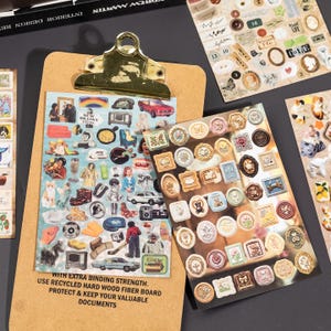 Puede incluir: Un portapapeles marr&oacute;n claro con un clip de metal dorado sostiene dos hojas de pegatinas. Una hoja presenta varias ilustraciones peque&ntilde;as y coloridas de objetos, personas y veh&iacute;culos. La otra hoja muestra numerosos adhesivos decorativos peque&ntilde;os, ornamentados y enmarcados.