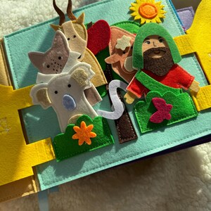 May include: A felt quiet book with a light blue background. The book features felt cutouts of animals, a man, a sun, and other elements. The animals include a goat, a fox, and a koala. The man has a green hood and a red shirt.