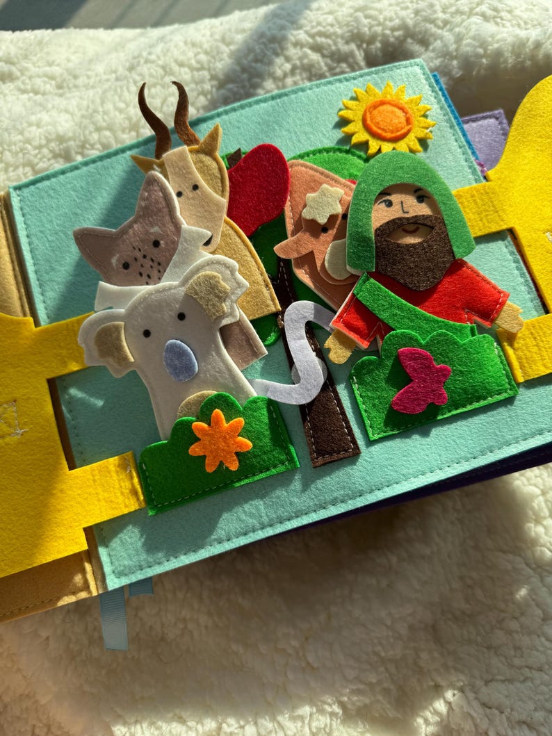 May include: A felt quiet book with a light blue background. The book features felt cutouts of animals, a man, a sun, and other elements. The animals include a goat, a fox, and a koala. The man has a green hood and a red shirt.