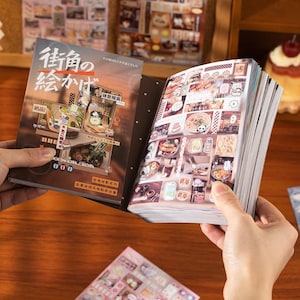 May include: A book with Japanese text and illustrations is open, revealing sticker sheets with food and character designs. The cover features a brown and beige design with Japanese text and illustrations. The book is held open by two hands.