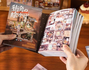 May include: A book with Japanese text and illustrations is open, revealing sticker sheets with food and character designs. The cover features a brown and beige design with Japanese text and illustrations. The book is held open by two hands.