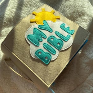 May include: A beige felt book with the words "MY BIBLE" in turquoise letters on a white cloud. A yellow sun is appliqued on the top. The book has a light beige handle and a blue ribbon bookmark.