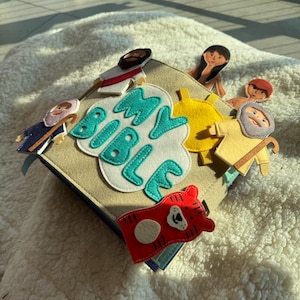 May include: A soft, beige fabric book with the words "MY BIBLE" in teal and white. The book is decorated with felt figures, including Jesus, a shepherd, and a red tiger. The book is open on a white, fluffy surface.