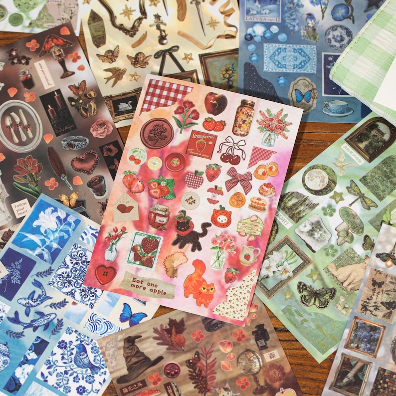 May include: A collection of decorative sticker sheets featuring various designs. The stickers include images of strawberries, cats, flowers, frames, and butterflies. The color palette includes red, pink, blue, and green.