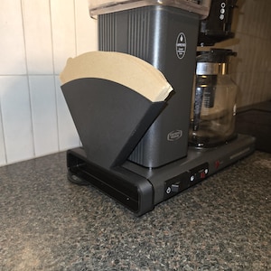 Può includere: Una macchina da caffè grigio scuro con un serbatoio dell'acqua trasparente e una caraffa di vetro. Un supporto nero con filtri per caffè di carta marrone è a sinistra. La macchina da caffè è su un piano di lavoro maculato.