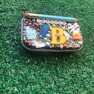 May include: A small black box with a glittery surface and a yellow letter "B" in the center. The box is decorated with various school supplies, including a pencil, paper clips, and a notebook. The word "TEACHER" is spelled out in white letters on the top of the box.