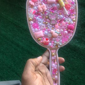 May include: A pink hand mirror decorated with various pink and white embellishments, including beads, bows, and charms. The mirror's surface is adorned with a crown, a lollipop, and a cupcake. The handle is also decorated with pink and gold accents.