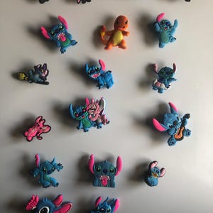 May include: A selection of colourful cartoon character charms, including Stitch and a Charmander figure. The charms are in various poses and designs, with vibrant blues, pinks, and oranges. These decorative charms are likely for shoes or accessories.