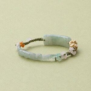 May include: A bracelet featuring a smooth, rectangular pale green jade piece, accented with small carved flower-shaped beads in light pink and orange, and textured round beige beads, all strung on a thin, olive green cord with knotted details.