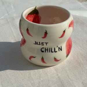 Può includere: Una tazza in ceramica bianca con una forma a clessidra unica. La tazza è decorata con illustrazioni di peperoncini rossi e la scritta "JUST CHILL'N" in lettere nere. L'interno della tazza è di colore rosa chiaro.
