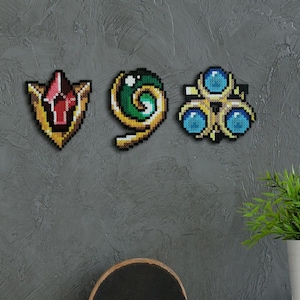 May include: Three pixel art designs: a shield with a red gem, a green and gold spiral, and a gold crown with blue gems. The designs are mounted on a grey wall, with a potted plant in the background.