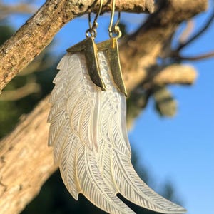 May include: A pair of earrings featuring a carved, iridescent wing design. The wings are white with detailed feather etching, suspended from gold-colored metal hooks. The earrings are displayed against a blurred background of a tree branch and blue sky.