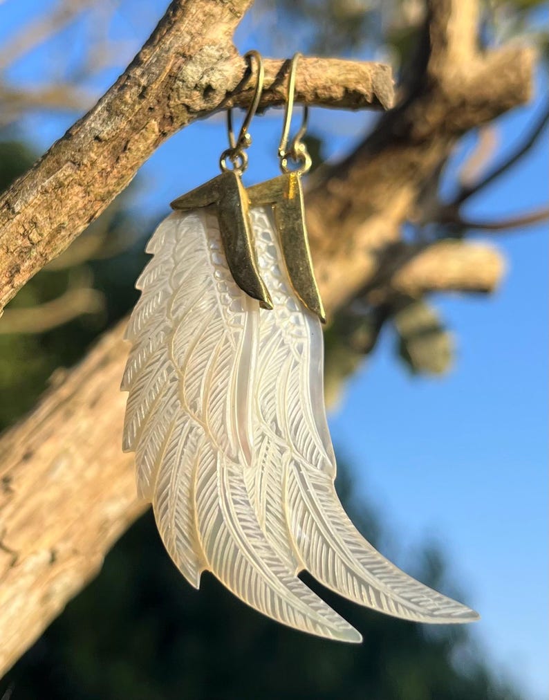 May include: A pair of earrings featuring a carved, iridescent wing design. The wings are white with detailed feather etching, suspended from gold-colored metal hooks. The earrings are displayed against a blurred background of a tree branch and blue sky.