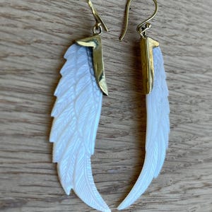 May include: A pair of earrings featuring a white, feather-shaped design with gold-colored accents. The earrings have a hook closure and are made of a pearlescent material, creating a shimmering effect. The earrings are displayed on a wooden surface.