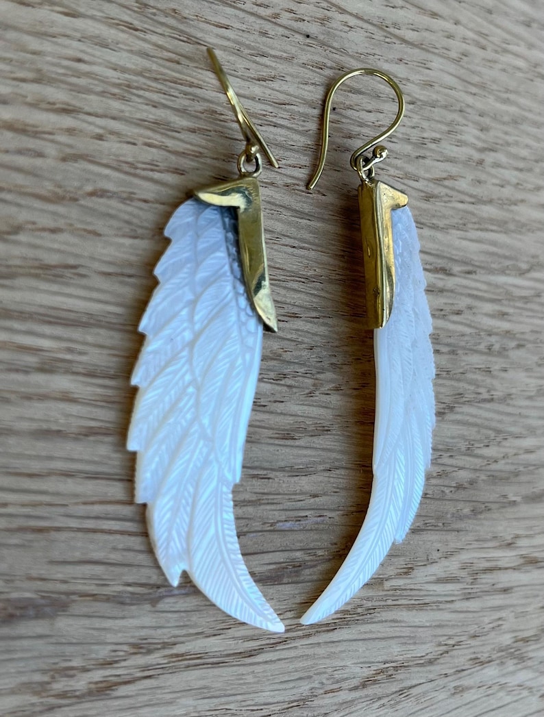 May include: A pair of earrings featuring a white, feather-shaped design with gold-colored accents. The earrings have a hook closure and are made of a pearlescent material, creating a shimmering effect. The earrings are displayed on a wooden surface.