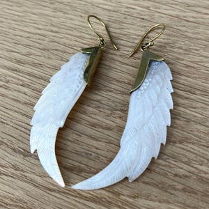 May include: A pair of earrings featuring a carved white feather design. The earrings have gold-colored hooks and accents. The feathers are detailed with carved lines, giving them a realistic appearance. The earrings are displayed on a wooden surface.