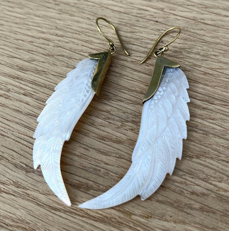May include: A pair of earrings featuring a carved white feather design. The earrings have gold-colored hooks and accents. The feathers are detailed with carved lines, giving them a realistic appearance. The earrings are displayed on a wooden surface.