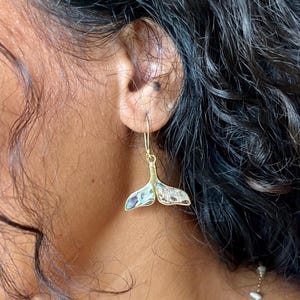 May include: A gold-toned dangle earring shaped like a whale tail, featuring an inlay of iridescent abalone shell with blue, green, and purple hues. The earring hangs from a simple gold-toned hook wire, showcasing a smooth metallic frame.